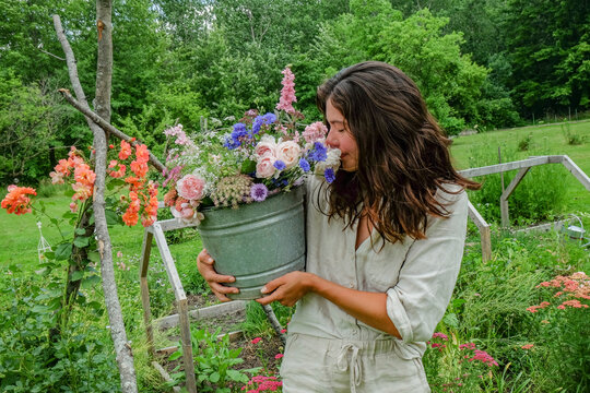 A woman in a light dress smiles as she holds a bucket brimming with colorful flowers in a lush garden.