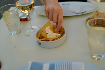 A person's hand picks a chip with dip from a bowl on a dining table with a glass of white wine and bread in the background.