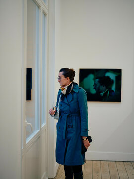 A woman in a teal coat with headphones stands contemplatively in an art gallery looking at a photograph.