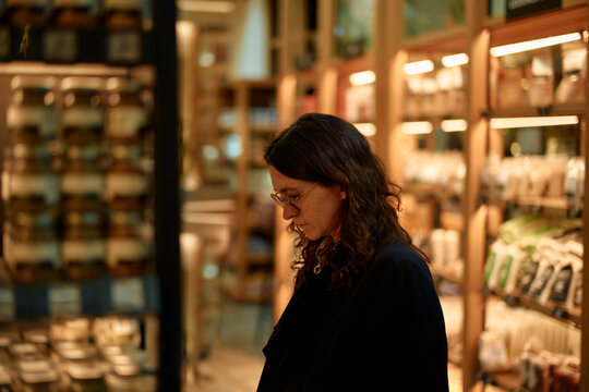 A woman in glasses peruses items in a warmly lit store with shelves stocked with goods in the background.
