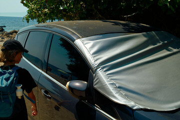 A child walks past a parked car with a sunshade on the windshield on a sunny beachside road.