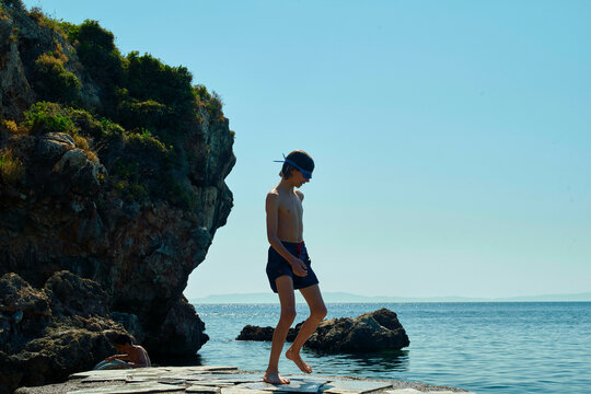 A person in swimwear walking on a rocky shore with clear blue water and a cliff in the background on a sunny day.