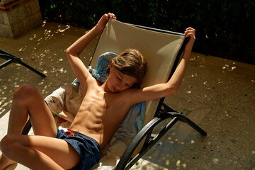 Young boy relaxing on a sun lounger in a sunlit courtyard, enjoying the warmth with eyes closed.