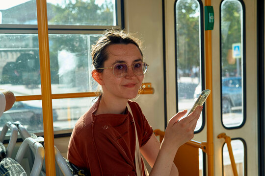 Smiling woman wearing sunglasses using a smartphone on a sunny tram ride.