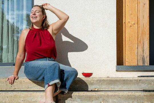 Smiling woman in a red top sitting on concrete steps next to a wooden door, enjoying the sun with a bowl of strawberries beside her.
