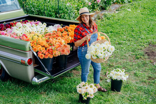 Woman in a straw hat and denim selling colorful flowers from the tailgate of a classic green pickup truck in a lush garden.