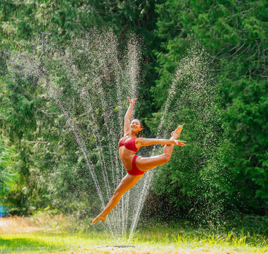 A joyful dancer performs an elegant leap amidst sparkling water jets in a sunlit green park.