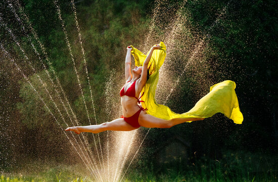 Woman in mid-air performing a split jump with yellow fabric against a backdrop of sunlit water sprinkles.