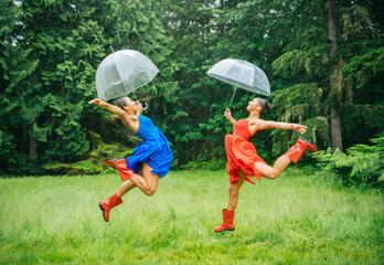 Two joyful women in colorful dresses and red boots leap up with transparent umbrellas on a grassy field with trees in the background.