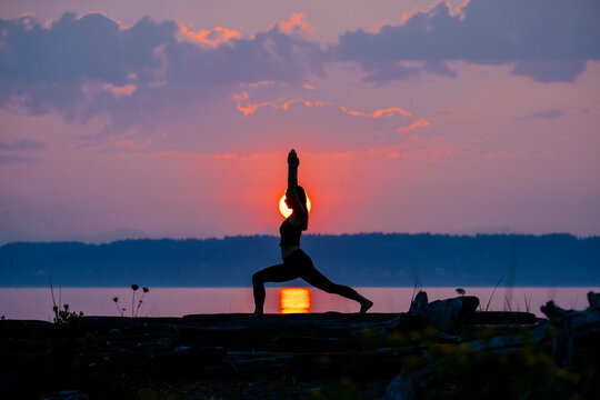 Silhouette of a person practicing yoga on a rock by a lake at sunset.