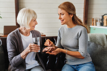 Senior woman taking pills from her daughter while holding glass with water.