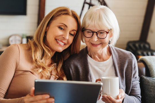 Cheerful senior woman with cup of tea and her daughter using tablet at home