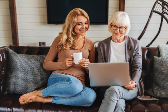 Smiling senior woman and her middle aged daughter sitting relax on sofa and drinking coffee in living room watching video on laptop together