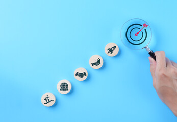 Business Goals and Target Success Concept. A hand using magnifying glass to focus on a target icon, business goals, and success. Icons of progress, teamwork, growth aligned, pathways to achievement.