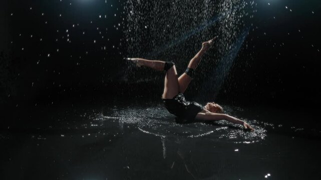 Graceful blonde woman in wet black dress sitting on floor and splashing water. Passion and emotions, beautiful slender lady playing with water in dark studio, slow motion, sexy female person moving