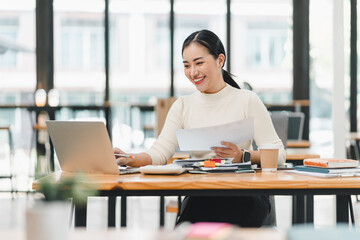 young woman is working at desk in modern office, smiling as she reviews documents on her laptop. bright environment enhances her focused and positive demeanor.