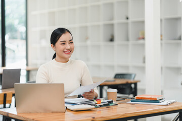woman is sitting at desk in modern office, smiling while reviewing documents. workspace is organized with laptop, stationery, and coffee cup, creating productive atmosphere.
