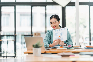 Fototapeta premium young woman is engaged in video conference, presenting data with chart in modern office setting. She appears focused and confident while using laptop and various documents on table.