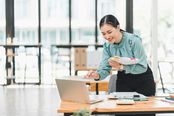 professional woman is smiling while working on laptop in modern office environment. She holds notebook and appears engaged in her tasks, showcasing positive and productive atmosphere.