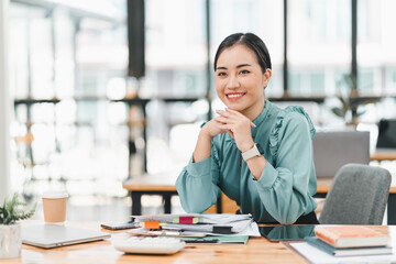 professional woman sits at desk in modern office, smiling confidently with her hands clasped. workspace is organized with documents, laptop, and coffee cup, creating productive atmosphere.