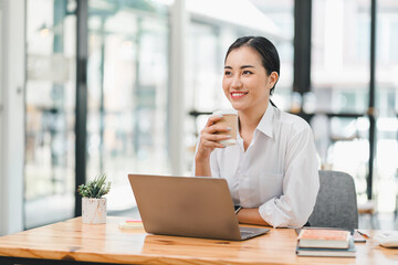 Fototapeta premium young woman is sitting at wooden table, smiling while holding coffee cup. She is working on laptop in bright, modern office space, surrounded by books and small plant.