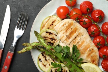 Delicious grilled vegetables and chicken breast served on black textured table, flat lay