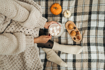 Woman in a knitted sweater sitting on a cozy plaid blanket, holding a cup of hot chocolate or cocoa drink with marshmallows. Hygge concept, top view