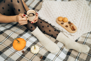 Woman in a black tights and warm socks sitting on a cozy plaid blanket, holding a cup of hot coffee drink