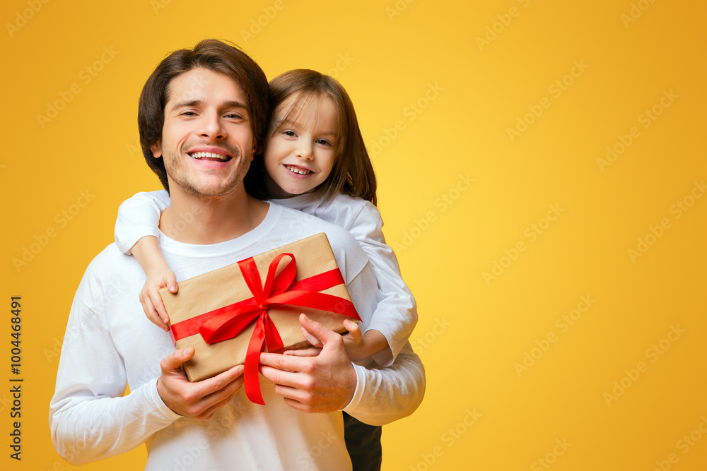 Wall mural Cheerful little daughter and father holding gift box, girl hugging dad from behind, Father Day concept, yellow background