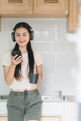 Enjoying moment of relaxation, young woman with long hair listens to music on her smartphone while holding cup in modern kitchen. Her casual outfit and headphones create cozy atmosphere.