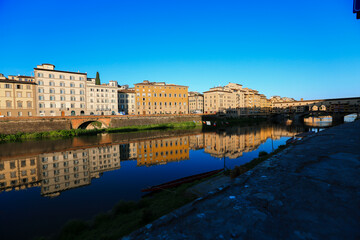 Fototapeta premium Reflections of Historic Florence along the Arno River The bright facades of buildings in Florence reflect perfectly on the calm waters of the Arno River, with the Ponte Vecchio in the distance.