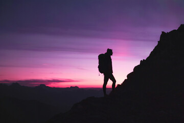 Fototapeta premium A silhouette of a person standing atop a rugged mountain peak, gazing into the horizon. The individual is equipped with a backpack, suggesting they might be on a hiking or trekking expedition. 