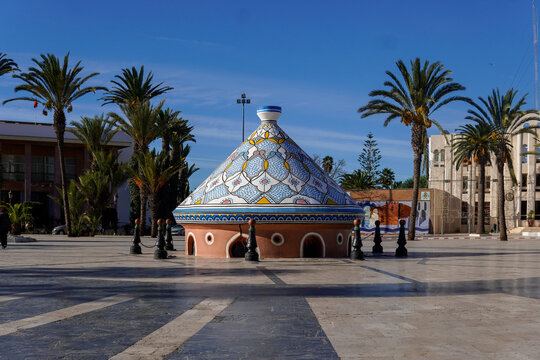 Colorful traditional Moroccan Biggest Tagine in a public square in Safi with palm trees and clear blue sky. A unique building with a conical tiled roof creates a striking landmark