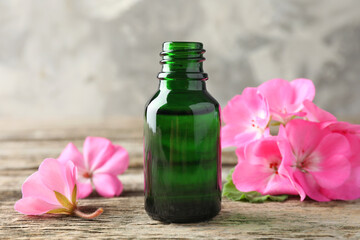 Bottle of geranium essential oil and beautiful flowers on wooden table, closeup