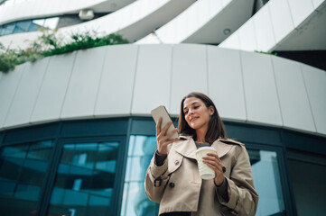 Businesswoman using smartphone and holding coffee outside office building