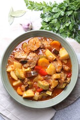 Delicious stew with vegetables in bowl, parsley, garlic and bay leaves on light grey table, top view