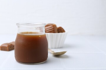 Tasty caramel sauce in jar, spoon and candies on white tiled table, closeup. Space for text