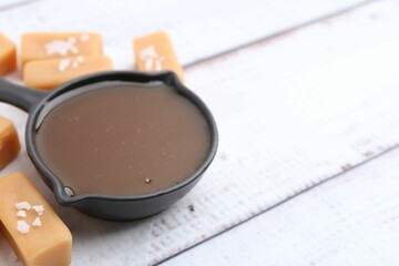 Tasty salted caramel in gravy boat and candies on white wooden table, closeup. Space for text