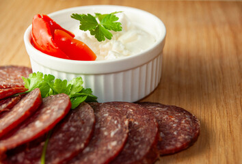 A fresh food arrangement with smoked sausage, and a bowl of fresh cow cheese with peppers, all displayed on a wooden cutting board.