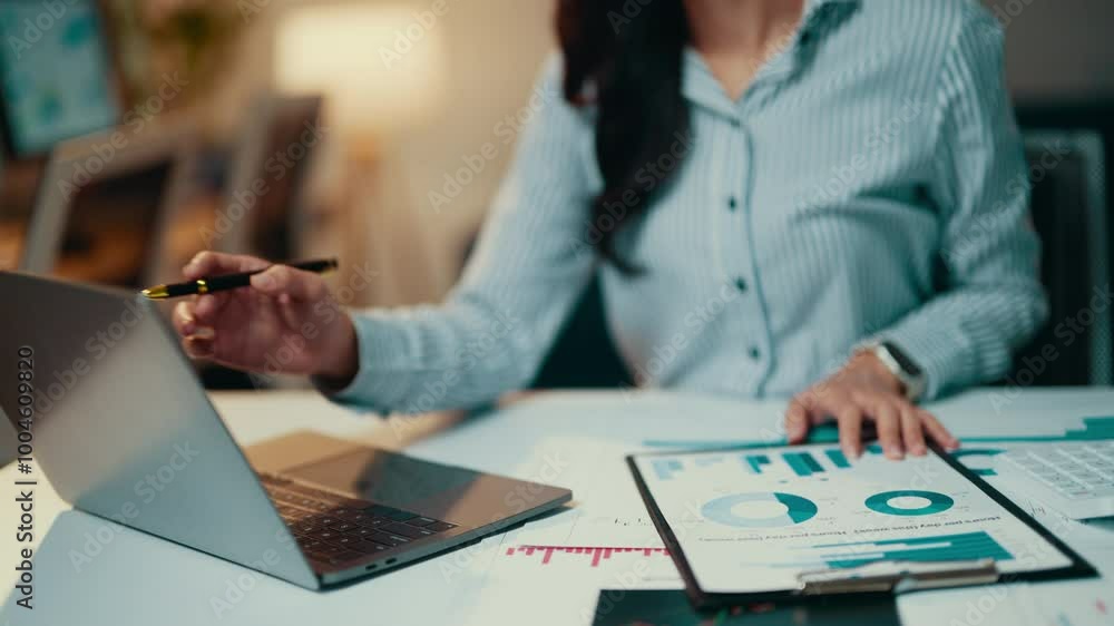 Poster businesswoman is sitting at her desk, working on a laptop and reviewing financial documents. she is 