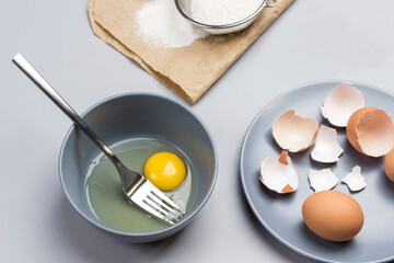 Broken egg and fork in gray bowl. Flour and sieve on paper. Chicken shells on gray plate.