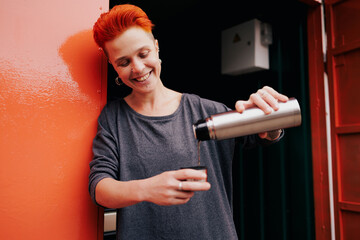 Smiling woman with vivid hair pouring drink from thermos near garage