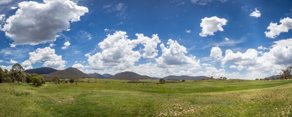 Panorama of Bogong Creek, where peaceful meadows stretch across Namadgi National Park. Lush green grass dotted with boulders and trees, all set against distant hills and a cloudy blue sky.