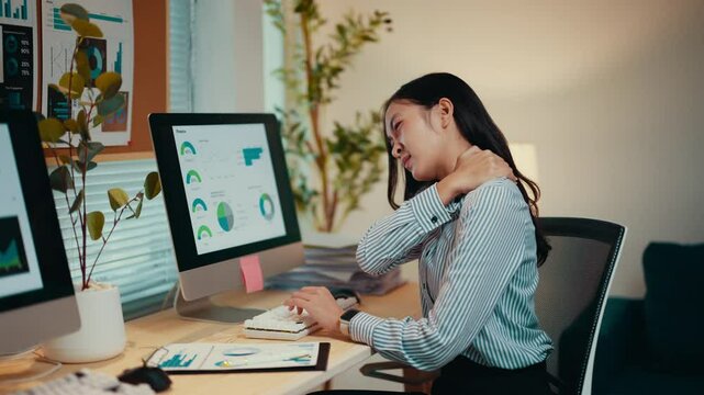 Asian businesswoman sitting at her desk suffers from neck pain while working on a computer. Stress and poor ergonomics contribute to her discomfort and fatigue in the office