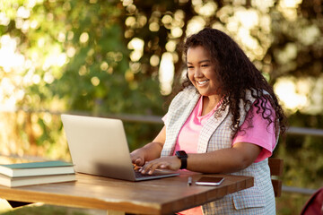 A woman with curly hair smiles while using her laptop at a wooden table surrounded by greenery. She has books and a phone nearby, enjoying a pleasant outdoor workspace in the afternoon.