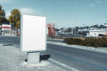 Empty urban billboard mockup on a modern city street. Blank advertising space in a sleek metal frame, standing on a cobblestone sidewalk with road, trees, and buildings in the background