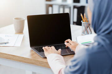 Unrecognizable muslim woman working on laptop with black screen, typing on keyboard, sitting in modern office, over shoulder view, mockup