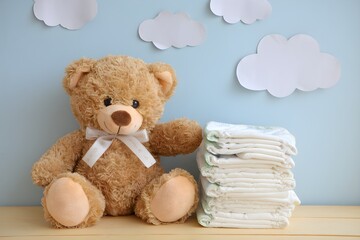 Brown teddy bear sits beside diapers on a wooden surface, cloud-decorated wall behind.