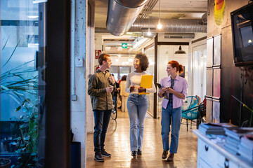 Obraz premium Three multiracial coworkers walking through coworking office. Group of young coworkers walking and talking in a brightly lit office hallway, carrying documents and tablets.