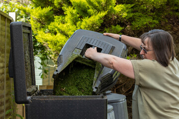 Middle age caucasian woman empties garden lawn mower grass clippings into compost hot bin composter